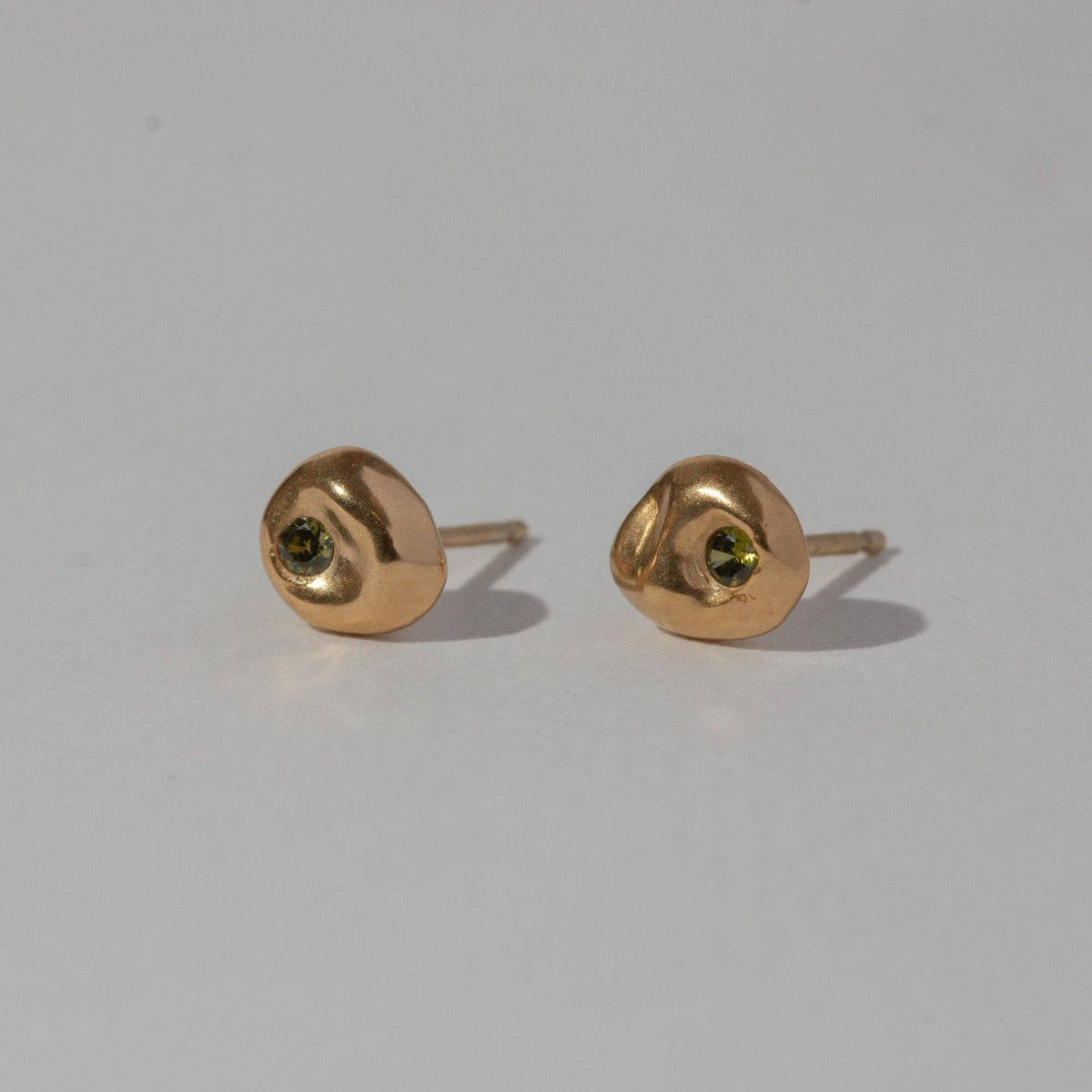 A pair of small gold stud earrings with a central green gemstone, displayed on a plain light gray background.
