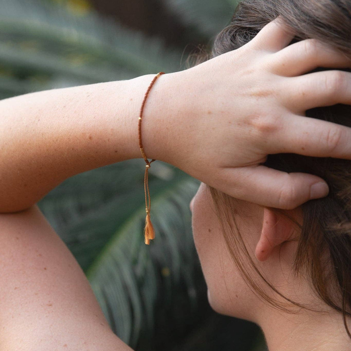 A close-up lifestyle shot of a woman from the side/back, wearing a delicate gold chain bracelet with a small gold tassel pendant on her wrist, set against a soft-focus background of green tropical leaves.