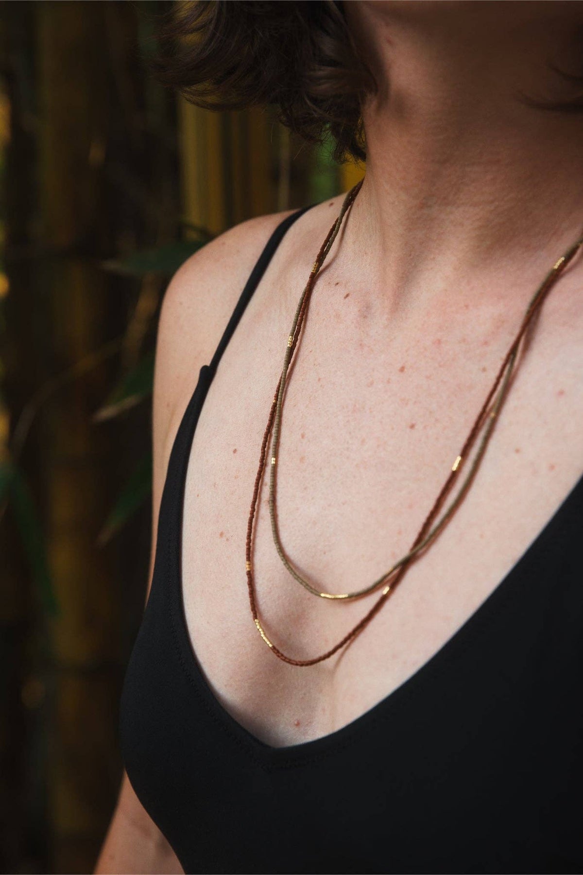 A close-up of a person wearing a layered necklace with thin gold and brown chains over a black camisole top, set against a dark, softly blurred background.