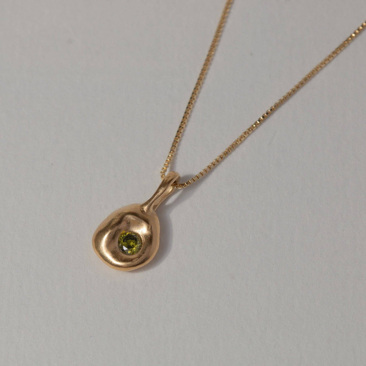 A gold chain necklace with a small, organic-shaped gold pendant featuring a central green gemstone, displayed on a plain light gray background.