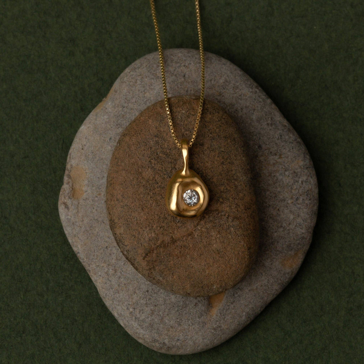 A gold pendant necklace with a small clear gemstone, displayed on a brown stone over a larger gray stone against a dark green background.