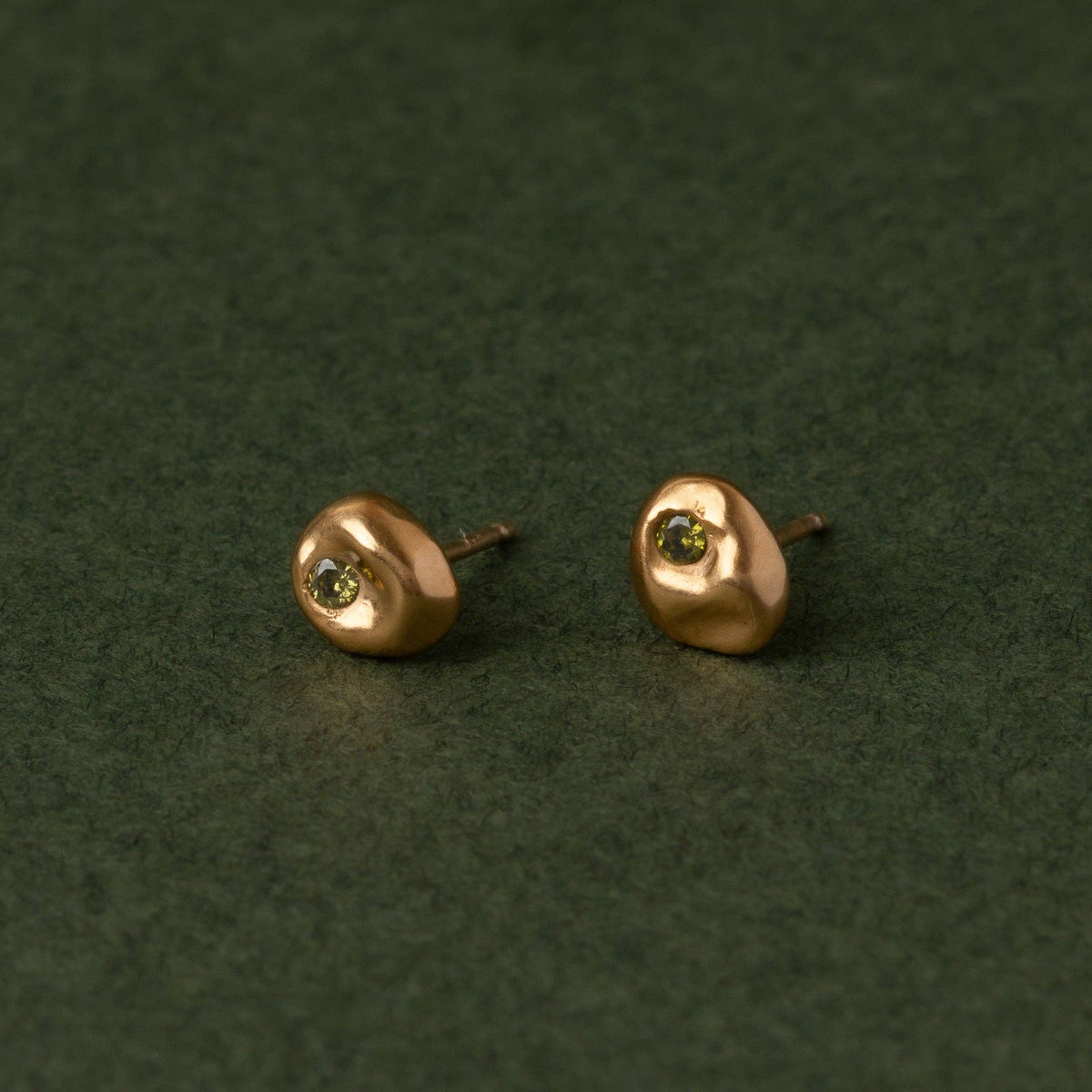 A pair of small gold stud earrings with a textured, organic shape and a tiny green gemstone in the center, displayed on a dark green textured background.