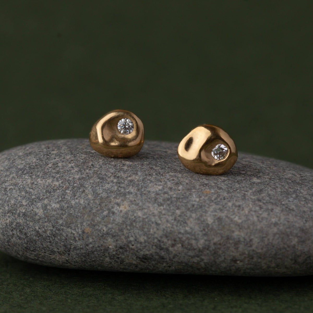 A pair of small gold stud earrings with a single clear gemstone, displayed on a smooth gray stone against a dark green background.