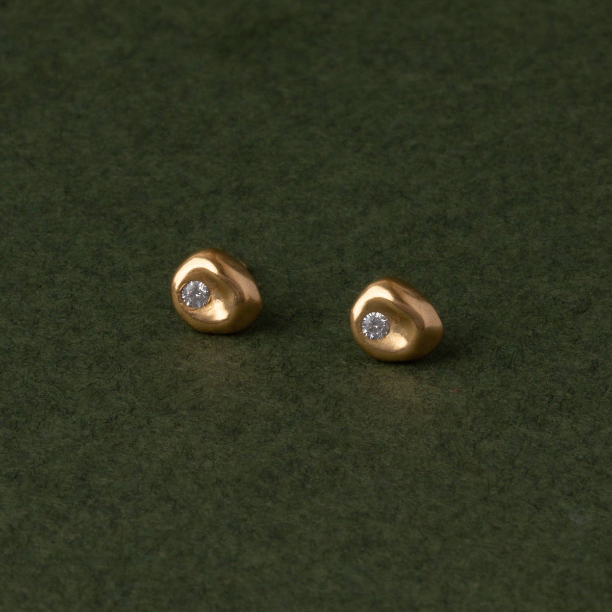 A pair of small gold stud earrings with a tiny clear gemstone in each, displayed on a dark green textured surface.