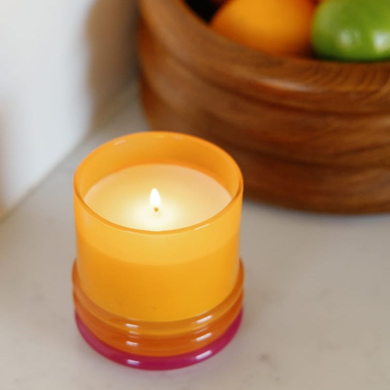 A lit orange candle in a translucent orange glass holder with a pink base, set on a light marble surface with a wooden bowl of fruit blurred in the background.